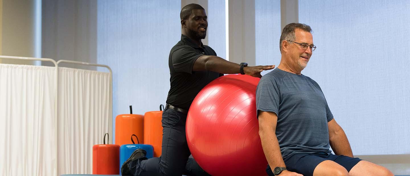 A male physical therapist helping a man with an exercise ball on a table in a fitness studio.